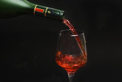 A red liquid is being poured from a green glass bottle into a transparent wine glass, with the liquid cascading down in a smooth stream. The backdrop is dark, emphasizing the rich color of the liquid.