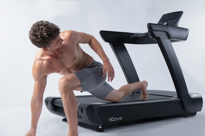 A shirtless man wearing gray shorts is positioned on the side of a treadmill, looking over his shoulder. He appears to be in mid-movement, possibly stretching or preparing to exercise. The treadmill is sleek and modern, set against a plain white background, which emphasizes the focus on the individual and the equipment.