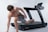 A shirtless man wearing gray shorts is positioned on the side of a treadmill, looking over his shoulder. He appears to be in mid-movement, possibly stretching or preparing to exercise. The treadmill is sleek and modern, set against a plain white background, which emphasizes the focus on the individual and the equipment.