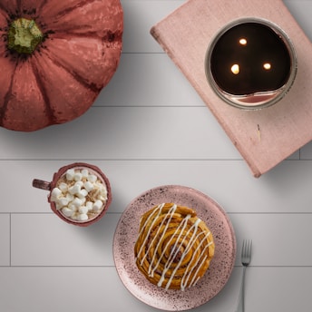 A cozy arrangement includes a large pumpkin, a hot beverage topped with marshmallows, a cinnamon roll on a speckled plate, a book beside a candle with a warm glow, all placed on a wooden surface.