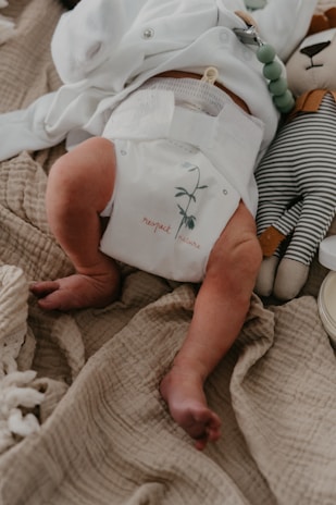A newborn baby is lying on a soft, textured beige blanket. The baby is wearing a white diaper with the words 'respect nature' and a small plant graphic. Next to the baby is a plush toy resembling an animal dressed in a striped outfit.