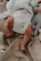 A newborn baby is lying on a soft, textured beige blanket. The baby is wearing a white diaper with the words 'respect nature' and a small plant graphic. Next to the baby is a plush toy resembling an animal dressed in a striped outfit.