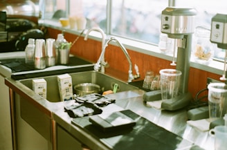 A kitchen or bar area featuring a stainless steel sink with a faucet, surrounded by various items including coffee cups, juice cartons, blenders, drink shakers, and glass containers. The counter has a clean and tidy appearance with some items placed on black mats, and natural light coming through the window.