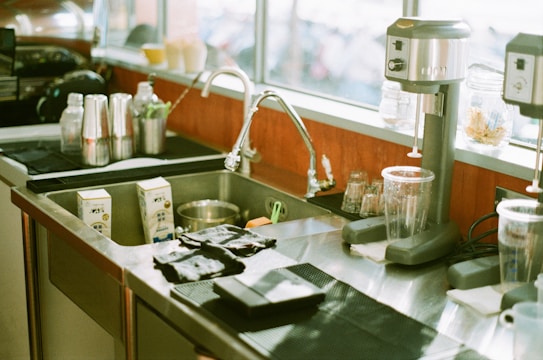 A kitchen or bar area featuring a stainless steel sink with a faucet, surrounded by various items including coffee cups, juice cartons, blenders, drink shakers, and glass containers. The counter has a clean and tidy appearance with some items placed on black mats, and natural light coming through the window.