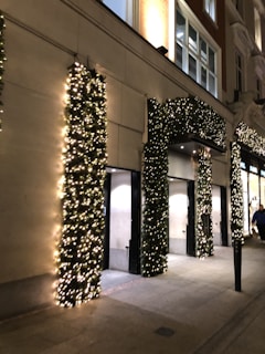 A building facade decorated with festive garlands wrapped around the columns and archways. The greenery is illuminated by numerous small white lights, creating a warm and inviting atmosphere. The sidewalk in front of the building is empty except for a person walking by, and the surroundings are softly lit by street lighting.