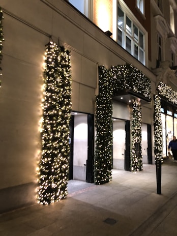 A building facade decorated with festive garlands wrapped around the columns and archways. The greenery is illuminated by numerous small white lights, creating a warm and inviting atmosphere. The sidewalk in front of the building is empty except for a person walking by, and the surroundings are softly lit by street lighting.
