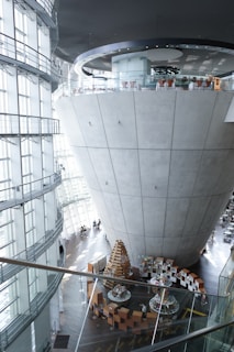 A large, modern architectural interior with a conical concrete structure at its center, topped with a circular balcony. The building features expansive glass windows allowing natural light to flood the space. There are people seated at tables and chairs around the base of the large structure and an arrangement of books and displays forming a circular pattern on the floor.