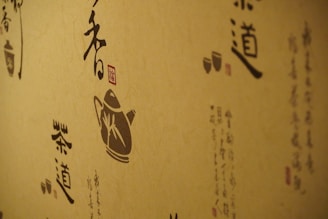 A wall decorated with artistic calligraphy and traditional illustrations, featuring a silhouette of a teapot and stylized Asian scripts. The designs are rendered in dark shades on a natural beige background, conveying a traditional and cultural aesthetic.