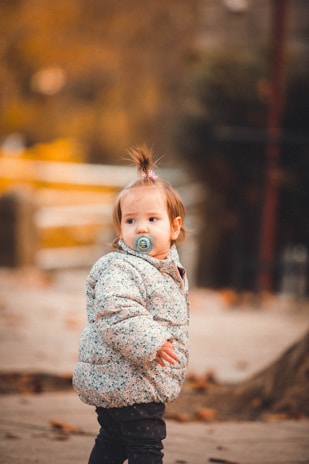 A young child with a pacifier stands outdoors, wearing a patterned winter jacket. The background is blurred with autumnal colors, and fallen leaves are scattered on the ground.