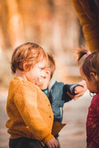 Three young children are interacting outdoors. One child in a yellow fuzzy jacket looks towards another child in a blue jacket, who appears to be holding the hand of an unseen adult. The third child, wearing a red outfit with a floral pattern and a ponytail, faces the first child. The background features warm, blurred autumnal tones.