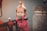 A young child with short, light brown hair tied in a small ponytail is holding onto the handlebars of a red and black tricycle. The child is wearing a pink jacket with patterns and has a pacifier in their mouth. The background is blurred, featuring a stone texture object to one side.