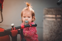A young child with short, light brown hair tied in a small ponytail is holding onto the handlebars of a red and black tricycle. The child is wearing a pink jacket with patterns and has a pacifier in their mouth. The background is blurred, featuring a stone texture object to one side.