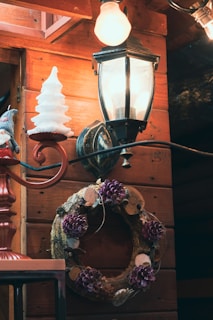 A rustic scene featuring a wooden wall adorned with a decorative wreath made of natural materials, including pinecones and purple flowers. A vintage-style wall lantern casts a warm glow, and there is a festive object resembling a white Christmas tree nearby.