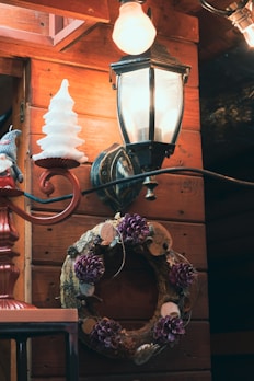 A rustic scene featuring a wooden wall adorned with a decorative wreath made of natural materials, including pinecones and purple flowers. A vintage-style wall lantern casts a warm glow, and there is a festive object resembling a white Christmas tree nearby.