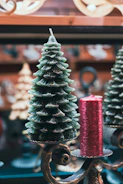 A decorative candle shaped like a Christmas tree is placed alongside a cylindrical red candle with a textured surface. Both are set on ornate metal holders with intricate scrollwork. The background features blurred holiday décor, creating a festive atmosphere.