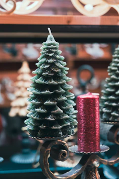 A decorative candle shaped like a Christmas tree is placed alongside a cylindrical red candle with a textured surface. Both are set on ornate metal holders with intricate scrollwork. The background features blurred holiday décor, creating a festive atmosphere.