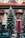 A decorative candle shaped like a Christmas tree is placed alongside a cylindrical red candle with a textured surface. Both are set on ornate metal holders with intricate scrollwork. The background features blurred holiday décor, creating a festive atmosphere.