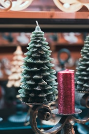 A decorative candle shaped like a Christmas tree is placed alongside a cylindrical red candle with a textured surface. Both are set on ornate metal holders with intricate scrollwork. The background features blurred holiday décor, creating a festive atmosphere.