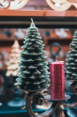 A decorative candle shaped like a Christmas tree is placed alongside a cylindrical red candle with a textured surface. Both are set on ornate metal holders with intricate scrollwork. The background features blurred holiday d&eacute;cor, creating a festive atmosphere.