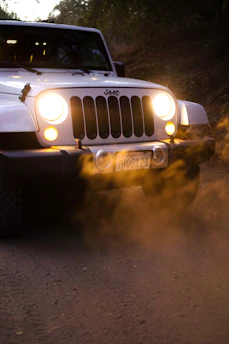 A white Jeep vehicle is driving on a dusty road with its headlights on, creating an effect of illuminated dust particles in the air. The image captures the front view of the vehicle, emphasizing its grill and circular headlights. The background appears to be a natural setting with some greenery.