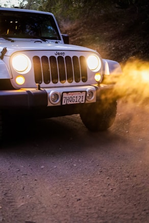 The image consists of a white Jeep with its headlights on, driving on a dirt road. The license plate reads 'California 7HQG121' and there is dust being kicked up from the tires. The setting appears to be outdoors, possibly in a mountainous or forested area.