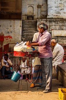 A vendor stands behind a small, red cart, preparing items for sale in an outdoor setting. He wears a checkered shirt and glasses. The cart contains items wrapped in plastic bags. Behind him, a few men are sitting and conversing, some dressed in traditional attire. The scene is set against a backdrop of an old, weathered building with stairs and faded wall art.
