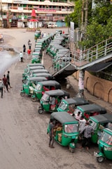 A line of green auto rickshaws is parked along a roadside near a pedestrian bridge. Several people are gathered around the vehicles, some loading goods or interacting with the drivers. Nearby buildings and street signage suggest a busy urban street corner.