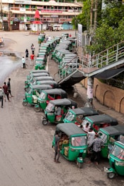 A line of green auto rickshaws is parked along a roadside near a pedestrian bridge. Several people are gathered around the vehicles, some loading goods or interacting with the drivers. Nearby buildings and street signage suggest a busy urban street corner.