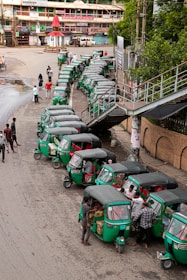 A line of green auto rickshaws is parked along a roadside near a pedestrian bridge. Several people are gathered around the vehicles, some loading goods or interacting with the drivers. Nearby buildings and street signage suggest a busy urban street corner.