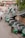 A line of green auto rickshaws is parked along a roadside near a pedestrian bridge. Several people are gathered around the vehicles, some loading goods or interacting with the drivers. Nearby buildings and street signage suggest a busy urban street corner.
