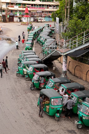 A line of green auto rickshaws is parked along a roadside near a pedestrian bridge. Several people are gathered around the vehicles, some loading goods or interacting with the drivers. Nearby buildings and street signage suggest a busy urban street corner.