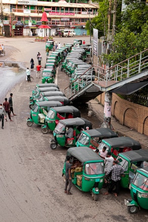 A line of green auto rickshaws is parked along a roadside near a pedestrian bridge. Several people are gathered around the vehicles, some loading goods or interacting with the drivers. Nearby buildings and street signage suggest a busy urban street corner.