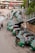 A line of green auto rickshaws is parked along a roadside near a pedestrian bridge. Several people are gathered around the vehicles, some loading goods or interacting with the drivers. Nearby buildings and street signage suggest a busy urban street corner.