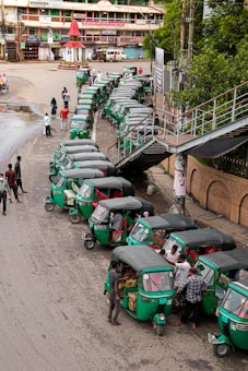 A line of green auto rickshaws is parked along a roadside near a pedestrian bridge. Several people are gathered around the vehicles, some loading goods or interacting with the drivers. Nearby buildings and street signage suggest a busy urban street corner.