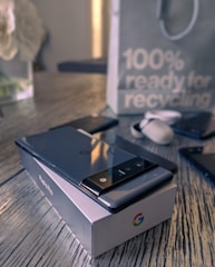 A smartphone with a sleek design rests on a branded box placed on a wooden surface. Nearby are earbuds and another phone. In the background, a paper bag features a message about recycling.