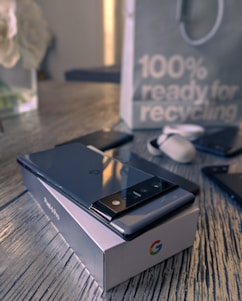 A smartphone with a sleek design rests on a branded box placed on a wooden surface. Nearby are earbuds and another phone. In the background, a paper bag features a message about recycling.