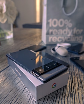A smartphone with a sleek design rests on a branded box placed on a wooden surface. Nearby are earbuds and another phone. In the background, a paper bag features a message about recycling.