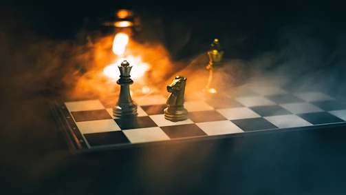 A chessboard is set with pieces under dramatic lighting. Visible pieces include a white queen and a gold knight, with a soft, warm light source in the background creating a smoky, atmospheric effect.