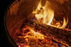 Copper smoker door open with glowing embers inside, surrounded by wood logs and smoke.