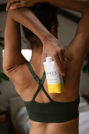 A person wearing a dark green sports bra holds a carton labeled as a vanilla bean flavored plant protein drink. Their arm is raised, and the carton is positioned against their back. The setting appears to be indoors, with soft lighting and blurred background elements.