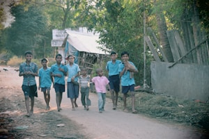 A group of children walk along a dirt path in a rural setting, two of them carrying small goats. They are dressed casually, and the background includes trees, a small house with a corrugated roof, and some wooden structures. The setting appears to be a village environment.