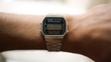 A digital wristwatch with a silver metal band is worn on a person's arm. The watch displays the time 04:20:00 on its digital screen. The watch case has a blue accent and is labeled as a Casio illuminator, water resistant with multiple buttons for adjustments.