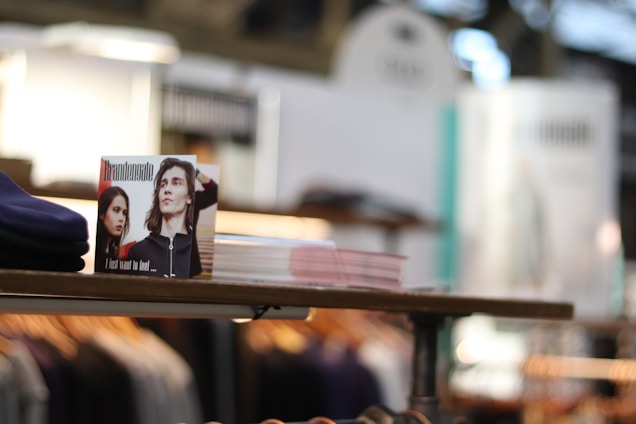 A stack of clothing displayed neatly on a wooden shelf at a store. In the foreground, there is a small advertisement card featuring two individuals with the text 'Brandengate' and 'I just want to feel...' printed on it. The background is blurred, showing racks of clothing and a bright, softly lit environment.