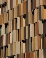 A facade of a building featuring numerous vertical panels in varying shades of brown and beige, arranged in an overlapping pattern. The panels have a perforated texture, allowing glimpse of the windows behind them.
