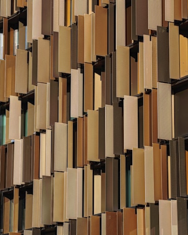 A facade of a building featuring numerous vertical panels in varying shades of brown and beige, arranged in an overlapping pattern. The panels have a perforated texture, allowing glimpse of the windows behind them.