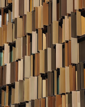 A facade of a building featuring numerous vertical panels in varying shades of brown and beige, arranged in an overlapping pattern. The panels have a perforated texture, allowing glimpse of the windows behind them.
