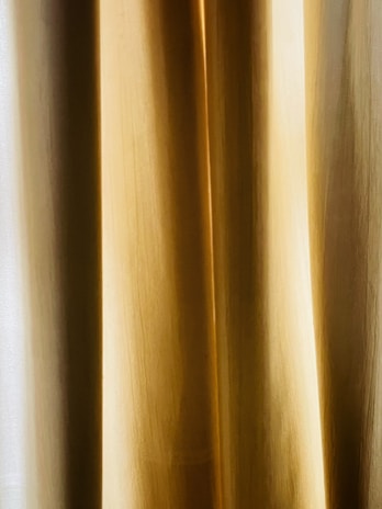 A close-up view of golden beige curtains with soft, vertical folds. The texture appears smooth, and the light creates subtle gradients.