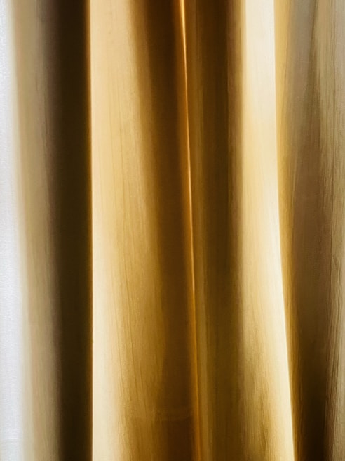 A close-up view of golden beige curtains with soft, vertical folds. The texture appears smooth, and the light creates subtle gradients.