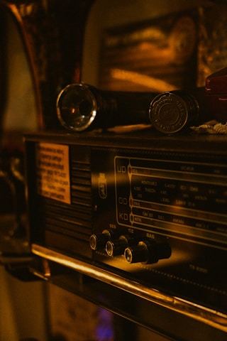 A vintage radio featuring knobs and a frequency display panel. On top of the radio, there is a flashlight and another small object. The lighting is warm, creating an antique and nostalgic atmosphere.