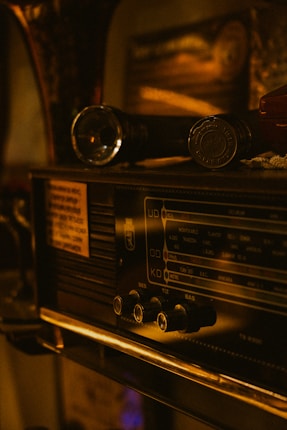 A vintage radio featuring knobs and a frequency display panel. On top of the radio, there is a flashlight and another small object. The lighting is warm, creating an antique and nostalgic atmosphere.